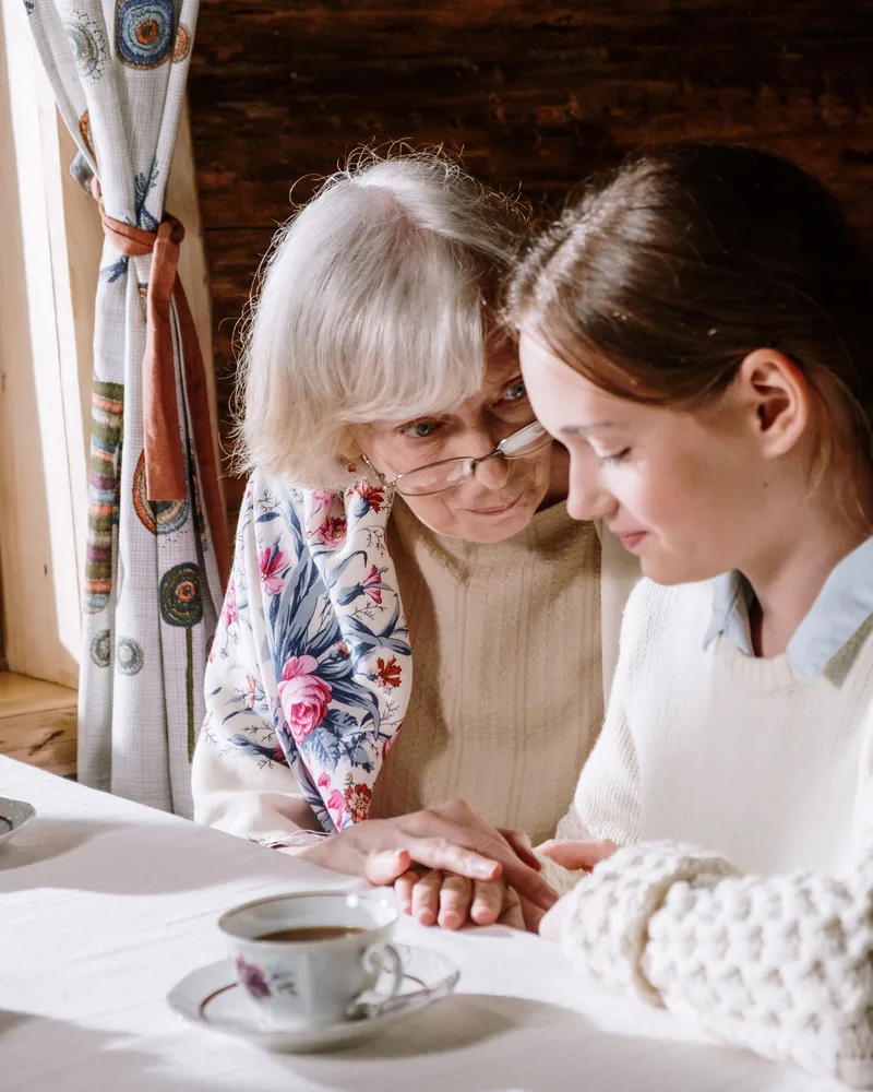 Senyva moteris ir jaunesnė moteris susikibusios rankomis prie stalo su arbatos puodeliu šiltoje jaukioje aplinkoje