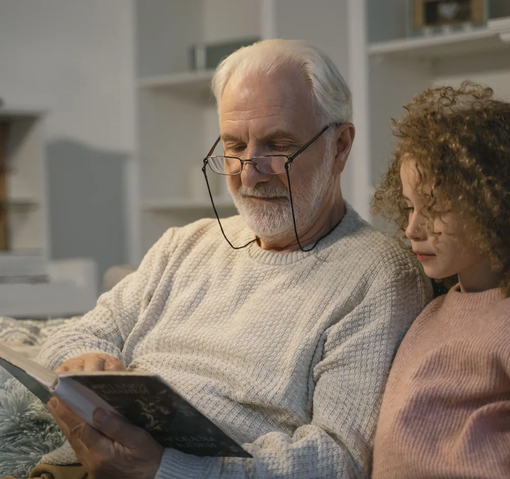Senelis su akiniais ir jo anūkė kartu skaito knygą jaukioje aplinkoje
